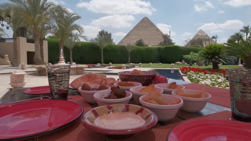 Egyptian food is on a table. Restaurant. Arabic cuisine. Pyramids of Giza is on the background. Luxury hotel terrace with palms and view on a Great Pyramids. Woman taking a photo.