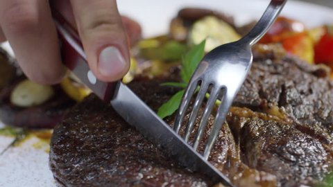 Closeup of male hands using knife and fork while cutting first bite from delicious beefsteak served with grilled vegetables
