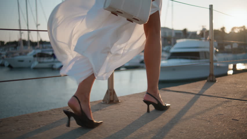 High-heels legs going on embankment summer windy evening close up. Unrecognizable stylish woman walking at waterfront in elegant white clothes. Unknown relaxed girl strolling pier near luxury yachts.