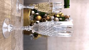 Vertical slow motion video of champagne being poured from a bottle into a glass with a Christmas tree in the background. Slow movement of liquids and bubbles in a champagne glass