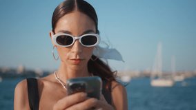 Trendy lady messaging at coast taking off sunglasses close up. Gorgeous happy woman smiling looking smartphone screen at sunny seashore. Beautiful girl reading message on cellphone standing beach. - Powered by Shutterstock - Get 15% off with code: PIKWIZARD15