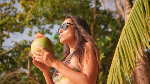 Attractive Mixed Race Young Tourist Girl Relaxing and Drinking Fresh Thai Coconut Water Cocktail at Tropical Beach. 4K, Slowmotion. Phuket, Thailand.