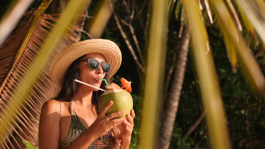Young Attractive Mixed Race Tourist Girl Drinking Fresh Thai Coconut Water at Tropical Beach. 4K, Slowmotion. Phuket, Thailand.