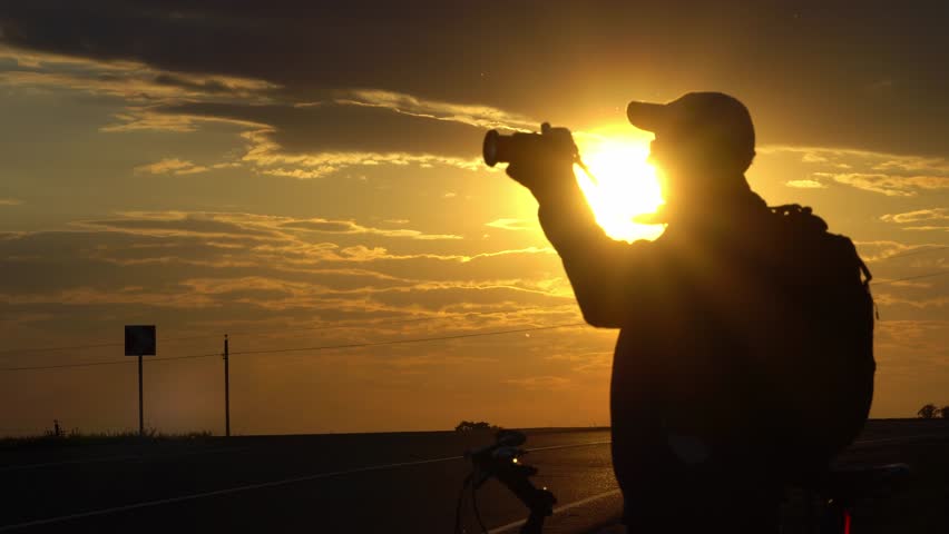 The silhouette of a guy with a camera against the sunset. A man is holding a camera in his hands. A photographer with a bicycle against the sky in the rays of the setting sun.