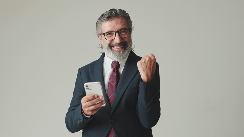 Businessman in glasses with gray hair, looking with smile at the mobile phone screen, getting good deal, winning, isolated on gray background in the studio | Shutterstock HD Video #3391126353