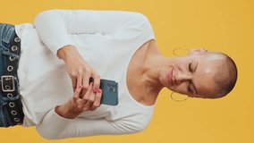 Vertical video, Smiling hairless woman, texting, communicating on social networks, mobile phone, isolated on yellow background in studio