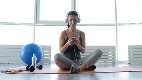 Delighted mood. Full length of positive young woman sitting on fitness mat while wearing earphones and listening to music