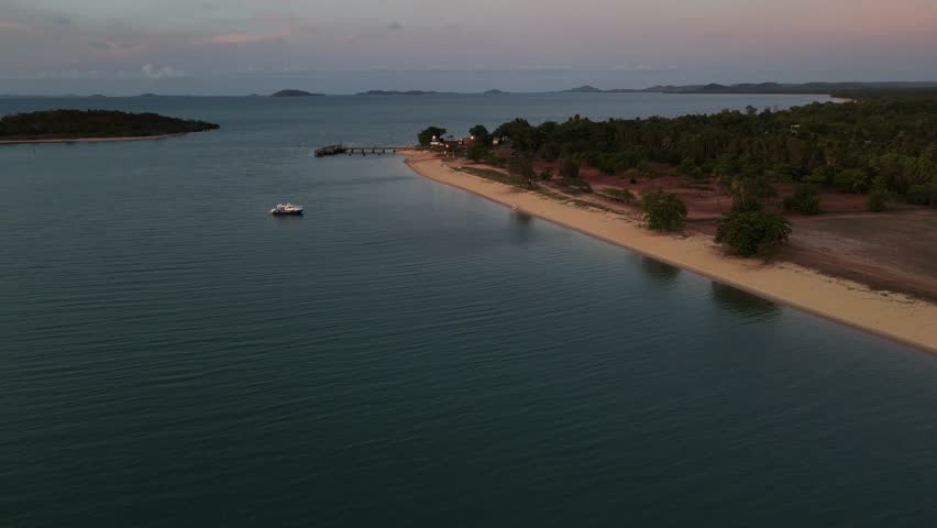 The last light of the day in remote north Queensland, Australia.  A large boat is anchored near shore, and there is a wharf that juts out from the sandy coastline.  Filmed in 4K.