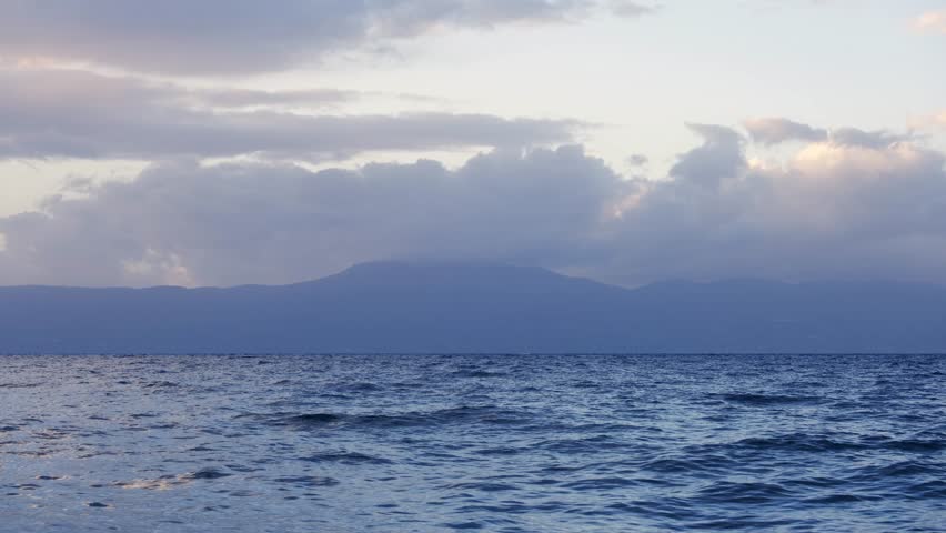 Pastel coloured tropical storm clouds moving above the wavy sea surface and the mountain range of Cres Island, Croatia. Magnificent landscape at the beach with cumulus clouds on a windy afternoon.