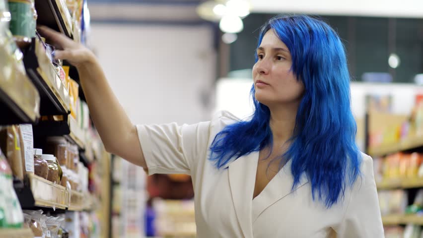 Young woman chooses package of brown sugar in supermarket. She takes plastic jar from shelf, looks at it, then puts it back and takes small bag. She puts it back on shelf as well
