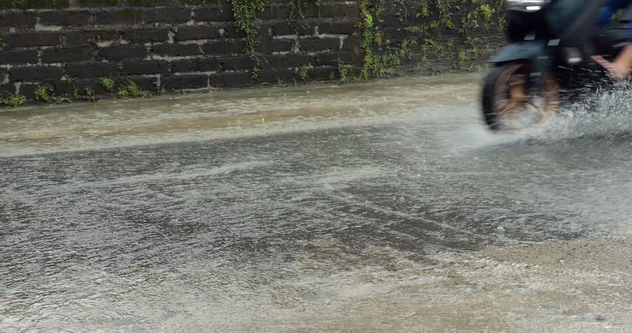 It's been raining all day, road has turned into small river, motorcycle rides through water with lots of splashing from wheels, slow motion footage. Low half side view, typical weather at rainy season