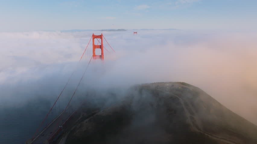 Aerial scenic white clouds covering tall red bridge on bright sunny evening. Cinematic dense thick fog at San Francisco. Flying above road and hills covered by cloudy fog to red Golden Gate Bridge 4K Royalty-Free Stock Footage #3391359427