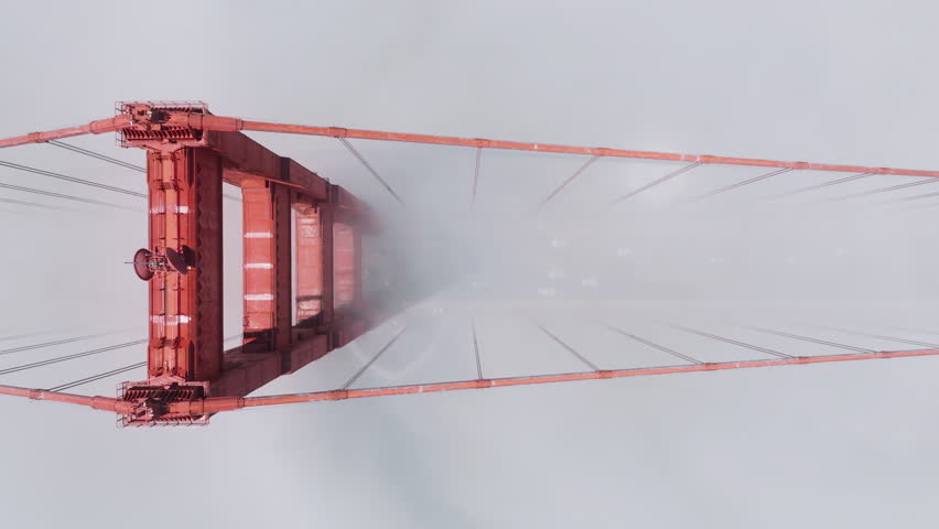Descending aerial shot overhead tall red bridge on bright sunny day. Sliding down above cars driving by road in dense thick fog, San Francisco. Traffic on Golden gate Bridge in scenic white cloud USA