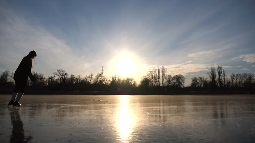 Young woman is skating on ice. Girl training alone at frozen lake or river. Lady improving his skills outdoor. Shining sun is reflecting on surface of ice. Active leisure at winter. Slow motion