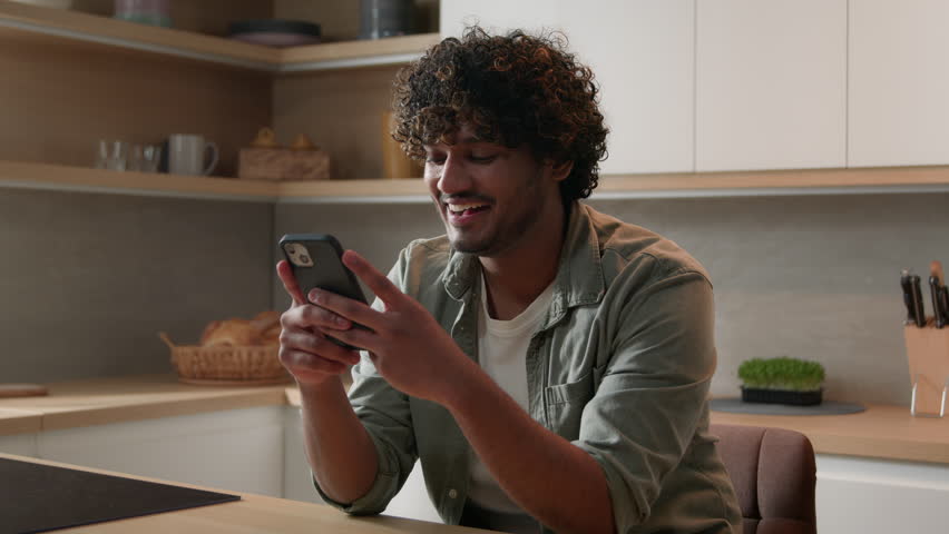 Moving shot carefree happy Indian man browsing smartphone laugh Arabian male smiling scrolling social media network app looking at mobile phone laughing smile enjoy funny video online at home kitchen