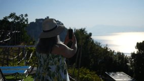 Happy female tourist in sunglasses and hat shooting video of Amalfi coast using smartphone, sharing photo on social media. Woman enjoying Italy vacation travel adventure. Shooting in slow motion.