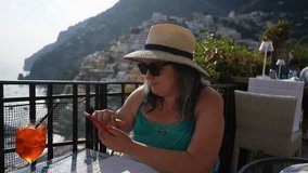 Female tourist in hat and sunglasses sitting at cafe table, using smartphone in Amalfi coast, taking photo, shooting video on background of beautiful seascape, on summer sunny day, slow motion.