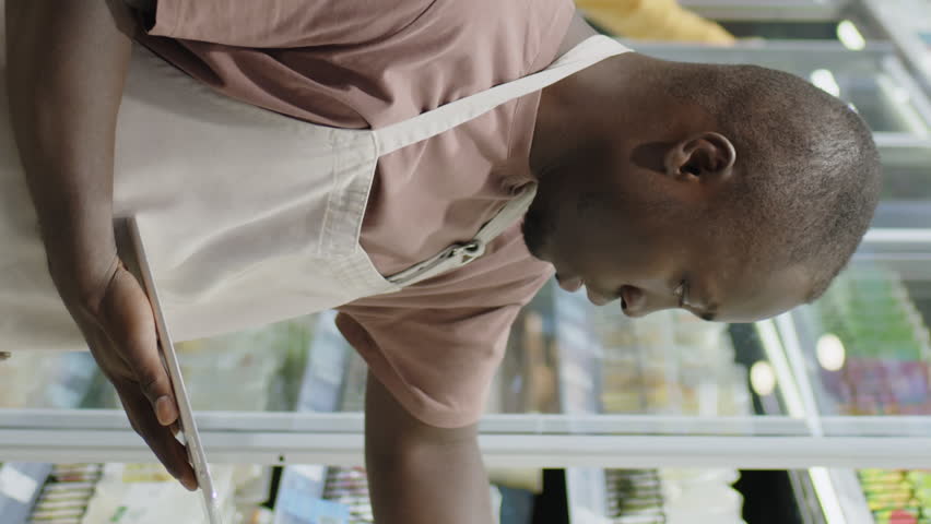 Vertical medium shot of young Black male supermarket worker in apron checking expiration data of packaged ready-made products and typing notes into digital tablet while standing in fridge aisle
