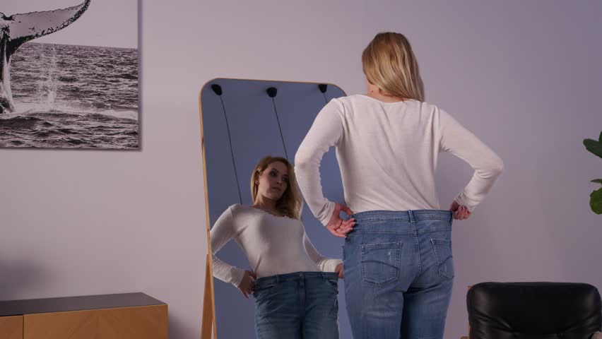 :Cheerful positive attractive middle aged woman in loose jeans, looking at reflection in mirror, showing achievement of successful weight loss diet and admiring body shapes indoors. Royalty-Free Stock Footage #3391746231