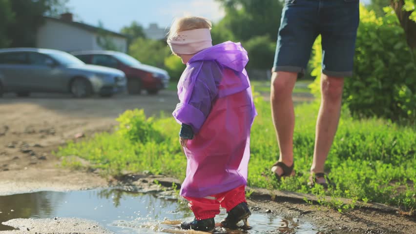A child aged two years in a purple raincoat and rubber boots runs through puddles in summer in sunny weather after rain. Slow mo