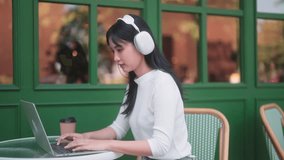 Asian woman is multitasking, typing on her laptop, collaborating with the team, and enjoying music through headphones. Amidst a Christmas-themed ambiance, balances work and holiday festivities