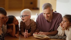 A male teacher with gray hair in a purple T-shirt reads a book to preschool children who lie on the floor on special pillows in a club for preparing children for school in a cozy room - Powered by Shutterstock - Get 15% off with code: PIKWIZARD15