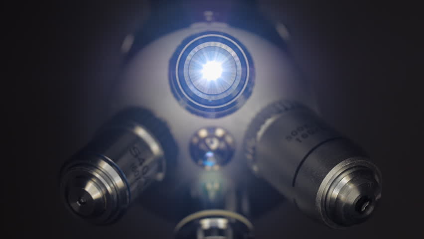 A hand rotates a block of microscope lenses one of them glows brightly on black background. Bottom view. Macro. Closeup. Shallow depth of field
