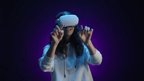 A happy girl in virtual reality glasses interacts with the virtual world using her fingers. She is in a room with a dark purple background illuminated by blue light. High quality 4k footage - Powered by Shutterstock - Get 15% off with code: PIKWIZARD15