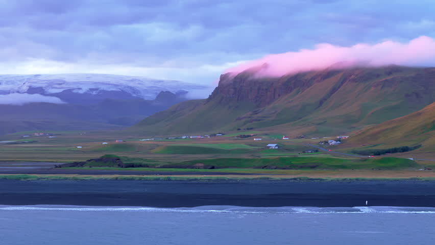 Astonishing landscape of icelandic black sand beach with pink clouds on tops of hills and green lowlands, snowy mountains and blinding rays of the sun. High quality 4k footage Royalty-Free Stock Footage #3392053075