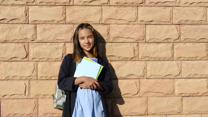 Portrait of a funny schoolgirl girl with a backpack looking at the camera and smiling while standing against a brick wall. Educational school program for children. Back to school.