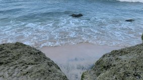 This video shows a beach scene with waves crashing onto the shore. The sand is covered in foam, and there are rocks on either side. The water is a deep blue color.
