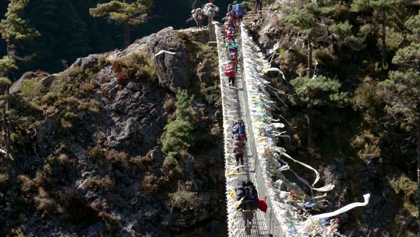 Trekking to Everest Base Camp. Tourists and sherpas with backpacks walk on Hillary suspension bridge through deep canyon of Himalayan mountains. Prayer flags and ribbons are fluttering in wind. Royalty-Free Stock Footage #3392104875