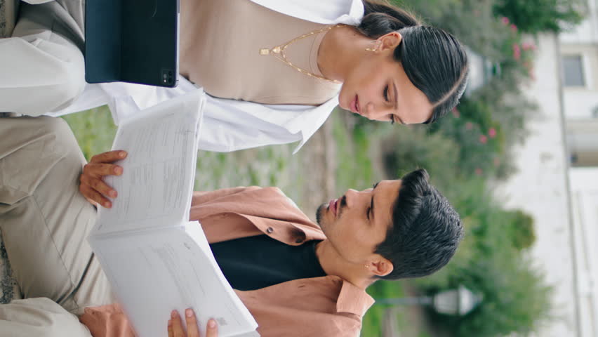 Serious couple working papers vertical. Latina business partners collaborating together using tablet computer on bench. Smart focused coworkers sitting at spring park nature. Busy teamwork concept