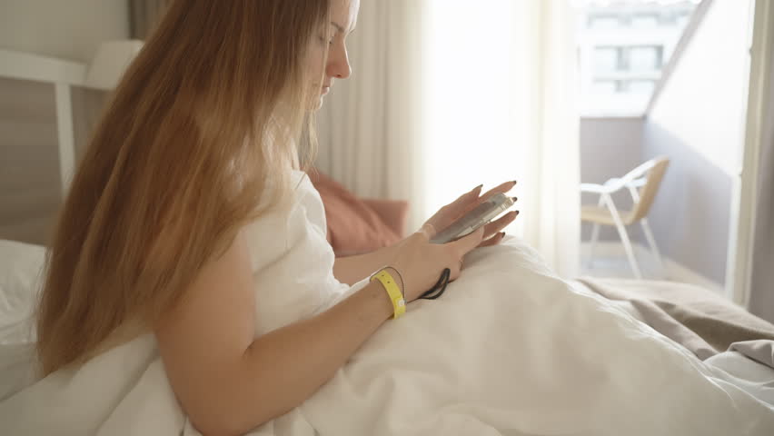 A Young Woman on a White Hotel Bed, Wearing a Yellow Guest Bracelet on Her Wrist. She is looking at something on her phone, possibly ordering room service. Sunbeams shine through the balcony door.