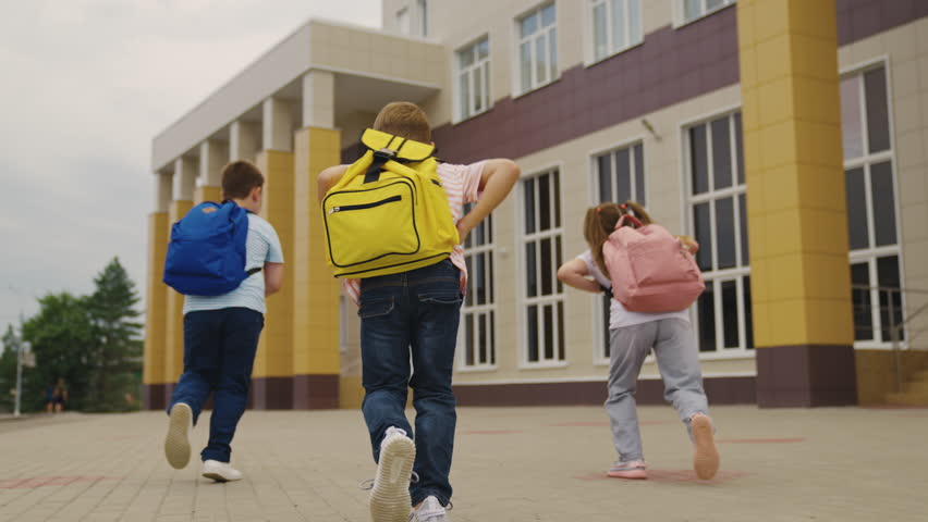 Playful children with schoolbags run together to school building backside view. Little students with backpacks hurry on school lessons. Active little children run to school in morning