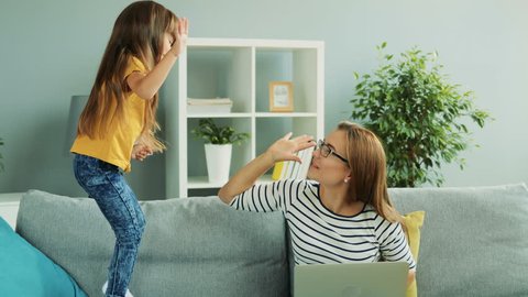 Attractive woman in glasses working on the laptop computer while her lovely nice daughter jumping on the couch next to her.   Inside  Epidemic, Self isolation, Home quarantine, pandemic, coronavirus  