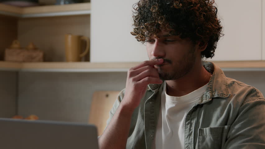 Close up Indian pensive man thinking computer problem solution typing laptop working online distant from home thoughtful Arabian male freelancer businessman think idea order food delivery in kitchen