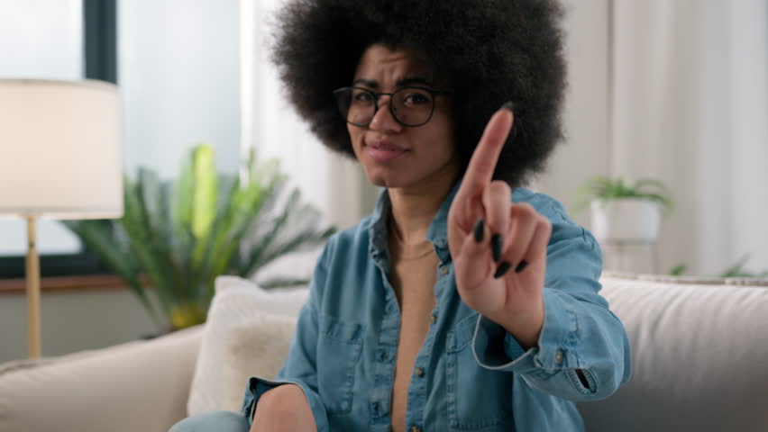 Serious confident ethnic girl African American woman at home sitting on couch sofa waving finger no not never gesture forbid showing negative answer attitude reject deny refuse reaction stop hand sign
