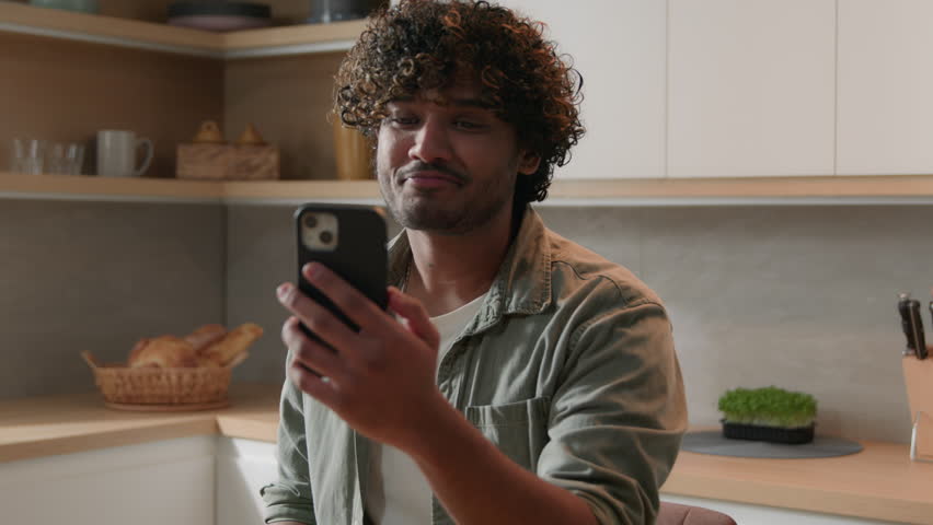 Happy smiling Indian man talking mobile phone virtual meeting online chat Arabian guy cheerful talk to online video call distant funny communication laughing speaking with friends at kitchen at home