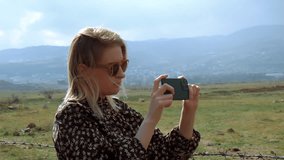 Young blonde woman holding smartphone and shooting video or taking shot. Real time middle shot. Outdoor nature travel concept. Memory and photography concept. High quality FullHD footage