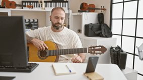Handsome young caucasian man musician engrossed in online classic guitar lesson at music studio, gracefully playing spanish acoustic instrument, eyes on smartphone screen