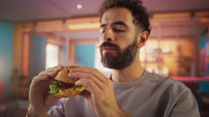 Fast Food Brand Commercial Concept: Slow Motion Portrait of a Young Stylish Man with a Beard Taking a Bite from a Delicious Hamburger, Looking at Camera and Smiling. Pure Enjoyment of Tasty Food Royalty-Free Stock Footage #3392681283