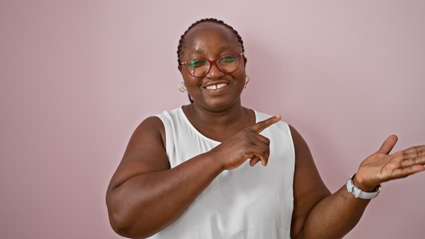 Joyful african american woman standing confidently, happily pointing and presenting over isolated pink background. adults expression of positive joy, showing copy space with fingers.