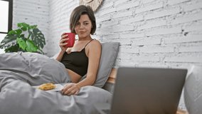 Happy young hispanic woman, confidently enjoying a joyous morning in her bedroom, sitting on bed, sipping morning coffee, while watching a movie on her laptop, as she relishes her breakfast