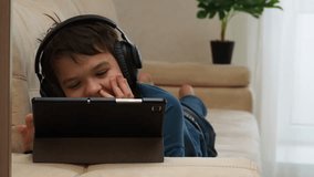 Smiling focused teenage school kid in big wireless headphones using tablet computer, relaxing on sofa at home, watching learning video, movie, attending online class, listening to music