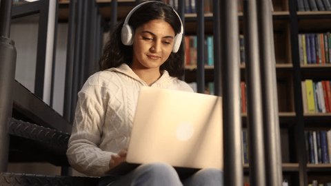 Portrait of pretty young woman student with headphones listening music while having distance remote education or work typing browsing scrolling on laptop computer at library or book store : vidéo de stock