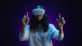 A happy girl in virtual reality glasses interacts with the virtual world using her fingers. She is in a room with a dark purple background illuminated by blue light. High quality 4k footage - Powered by Shutterstock - Get 15% off with code: PIKWIZARD15
