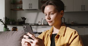 Happy woman at home, sitting on sofa and using her smartphone to chat and browse internet, close up. Enjoying her free time and casual lifestyle, pleasant ambience of modern living room. Cinematic AD