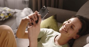 Young woman reclined on sofa, in sunshine and communicate with somebody online using smartphone. Smiling and laughing, enjoying free time, in psychological balance comfort. Cinematic advertising.