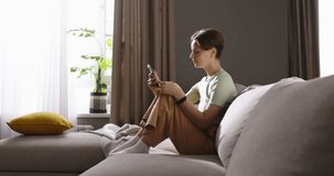 Attractive smiling young lady sits on sofa, savoring her free time with smartphone, texting with loved one boyfriend or friends. Peaceful moments, tranquility and happiness. Cinematic advertising